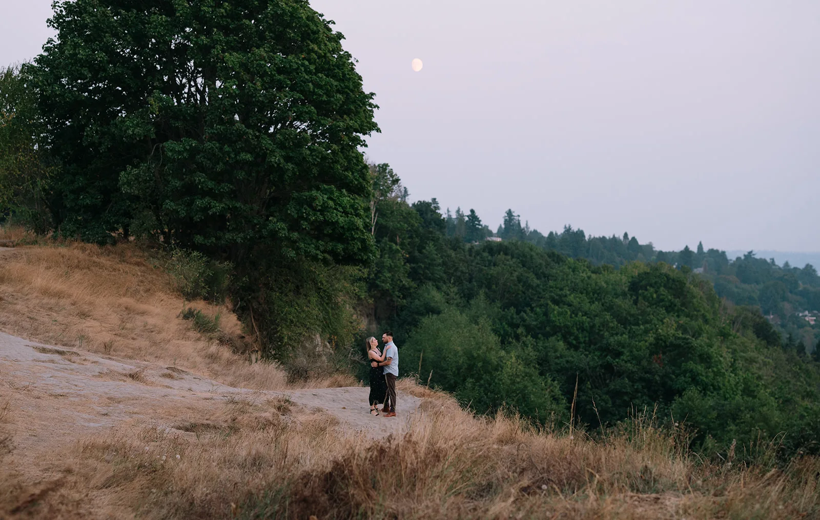 Couple standing by the shore at Discovery Park, with the woman wearing a green dress and the man embracing her.