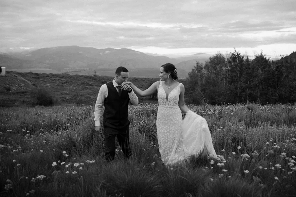 Black and white photo of a bride in a lace dress and a groom in a vest, sharing a gentle moment in a wildflower field with mountains in the background.