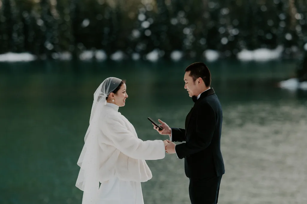Bride and groom holding hands and exchanging vows in a snowy elopement ceremony at Gold Creek Pond, with the forest's winter tranquility enveloping their special moment.