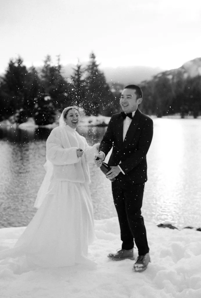 Bride and groom in a joyful champagne toast, creating a spray of sparkling droplets against the snowy landscape of Gold Creek Pond, encapsulating the happiness of their winter elopement.