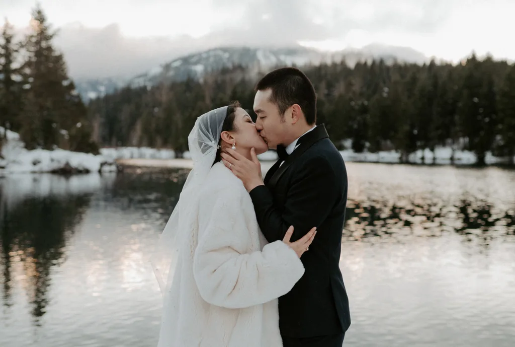 Newlyweds share a romantic first kiss by the reflective waters of Gold Creek Pond, with snowy mountains in the background, encapsulating the beauty of their winter mountain elopement.