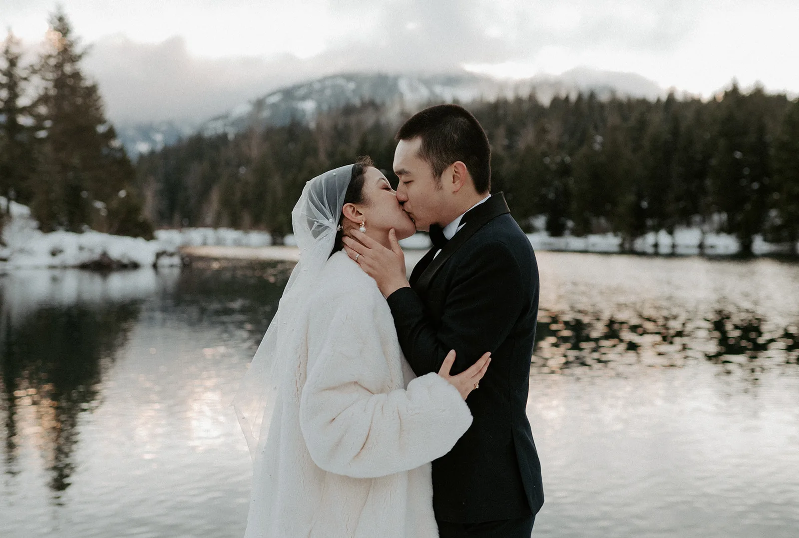 Newlyweds share a romantic kiss by the reflective waters of a snowy mountain lake.