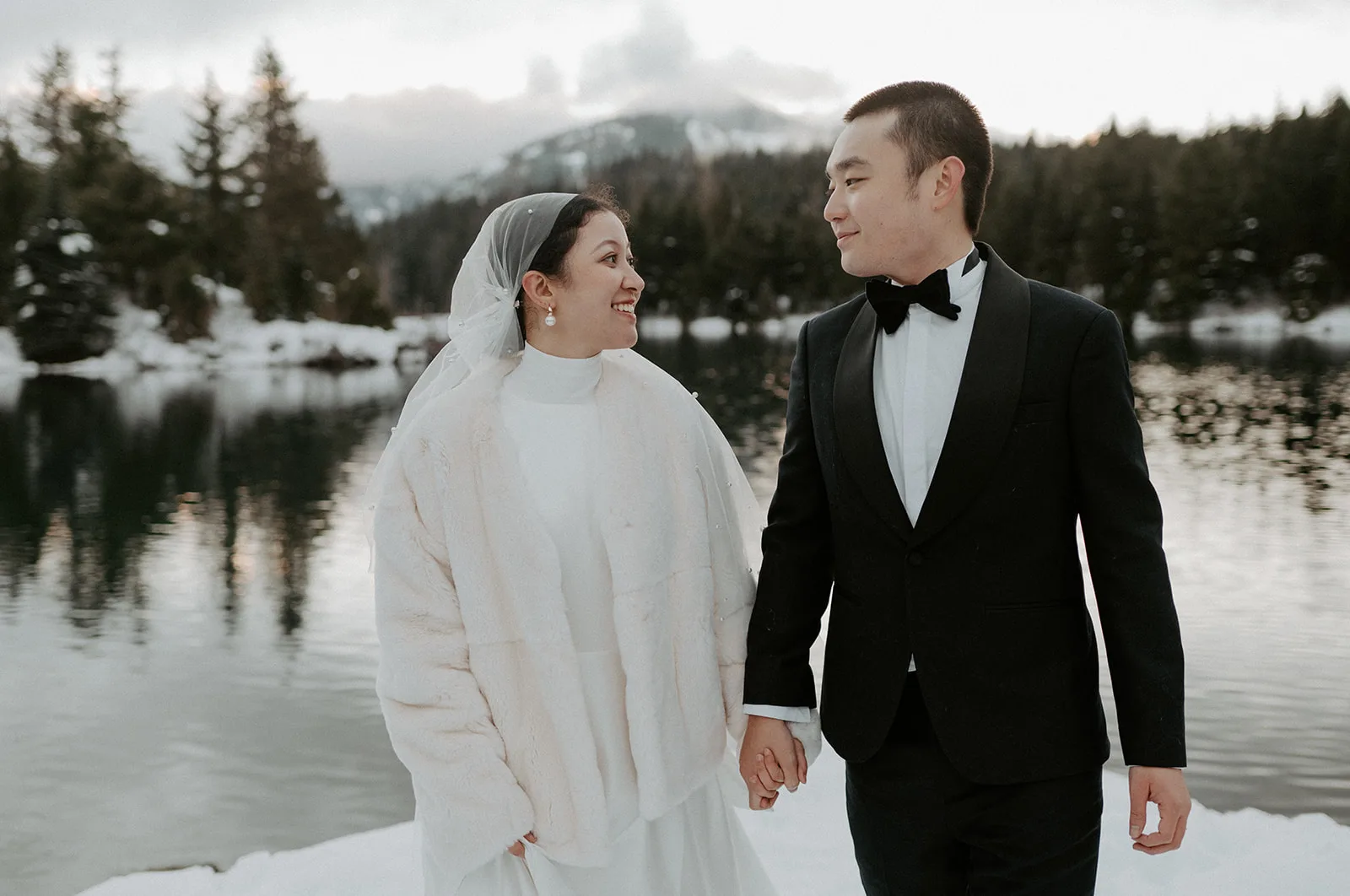 A beaming bride and groom walk hand in hand among a snowy lake landscape.