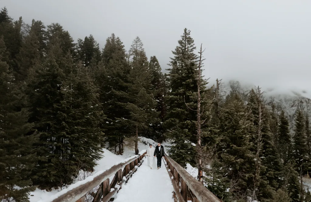 Bride and groom walk hand in hand across a snow-covered bridge at Gold Creek Pond, flanked by tall pine trees, encapsulating the serene and intimate essence of a snowy mountain elopement.