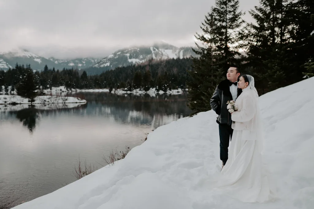 Bride and groom stand together, enveloped in the quiet beauty of Gold Creek Pond's snowy scenery, with mountain reflections in the calm waters creating a serene moment during their winter elopement.