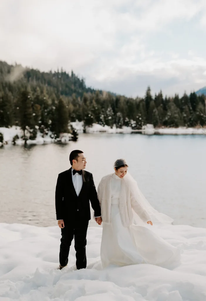Eloping couple holding hands and walking through the snow at Gold Creek Pond, with the bride in a white gown and veil, and the groom in a classic tuxedo.