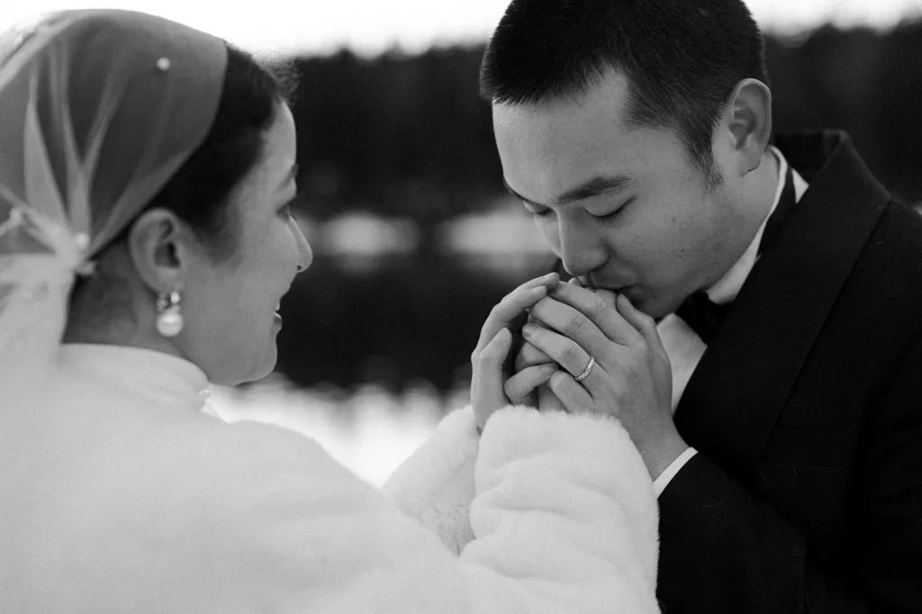 A poignant black and white photo of a groom kissing the bride's hands, conveying deep affection during their snowy elopement at Gold Creek Pond, with winter's gentle touch surrounding them.
