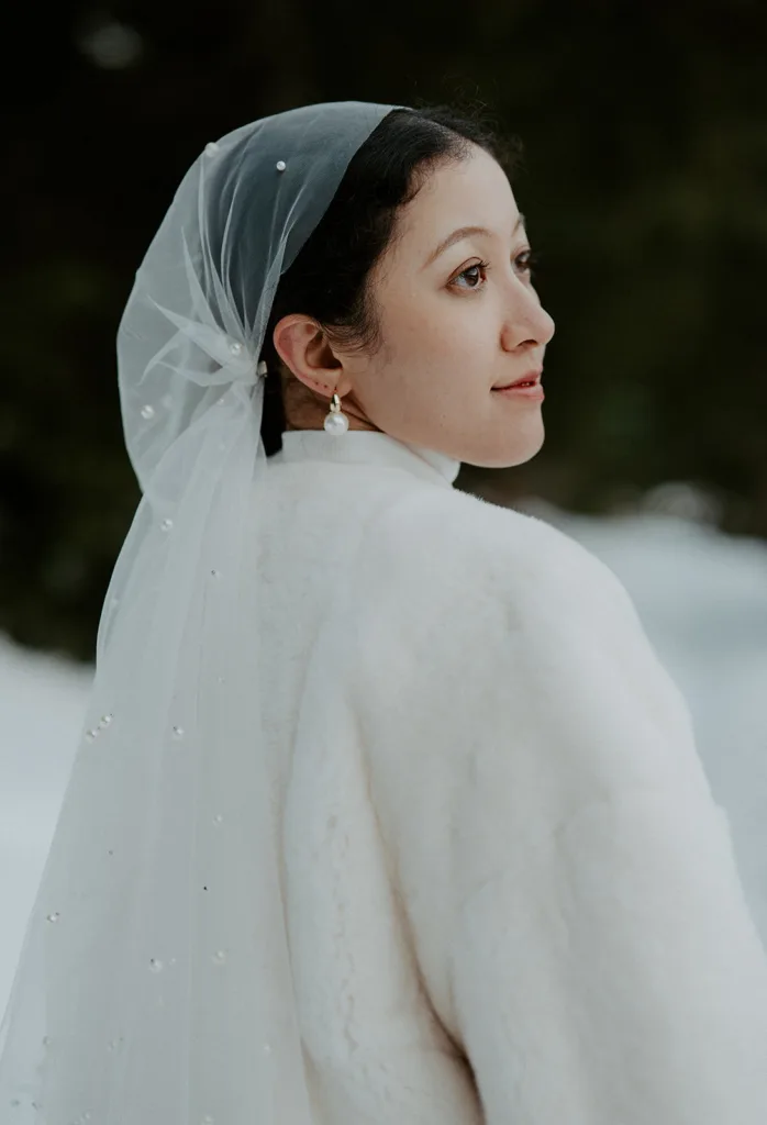 Close-up of a serene bride in a fur shawl and pearl earrings, her delicate veil adorned with pearls, set against the soft, white landscape of a snowy elopement at Gold Creek Pond.