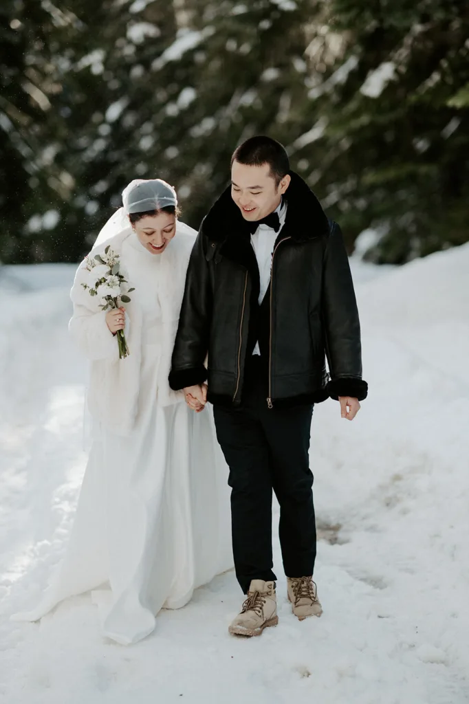 A bride and groom walk hand-in-hand through a snowy trail, sharing laughter in a quiet forest setting, symbolizing the intimacy and adventure of a mountain elopement.