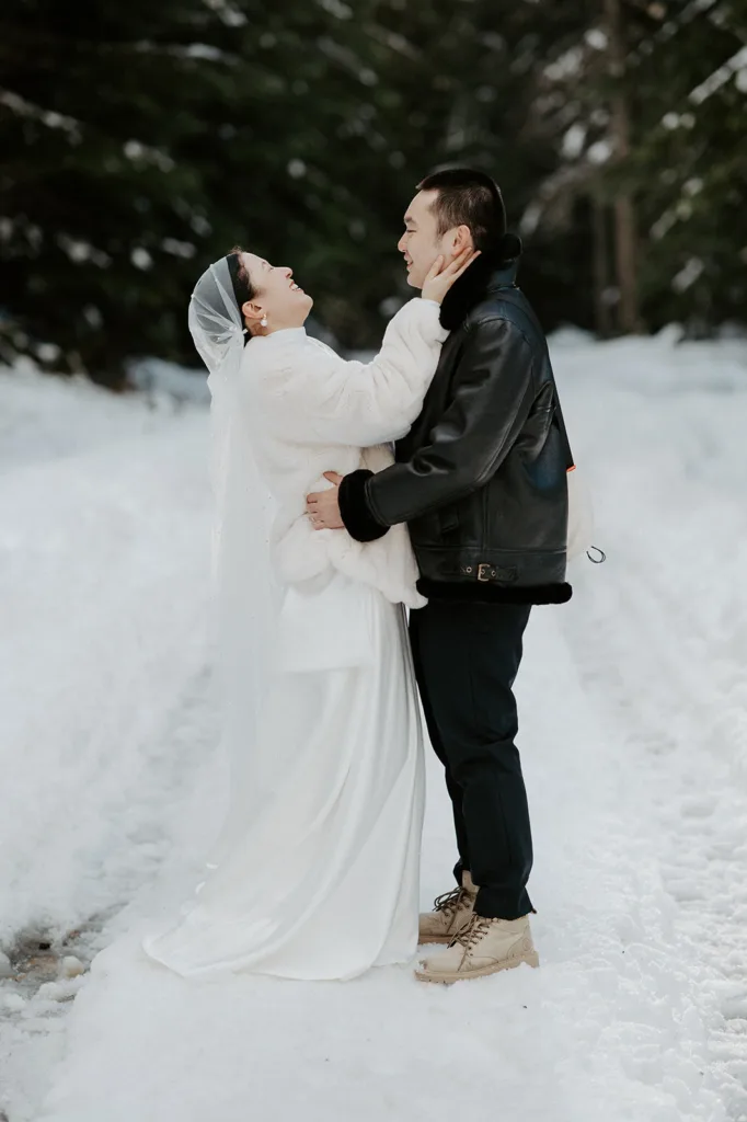 Bride in a white fur coat and groom in a leather jacket share a joyful moment during their snowy mountain elopement, surrounded by a peaceful winter forest setting.