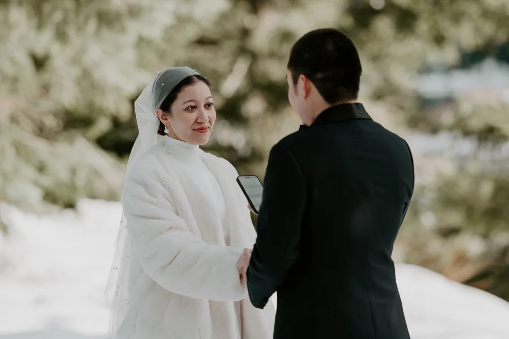 Bride in a white fur coat exchanging vows with her groom in an intimate snowy elopement at Gold Creek Pond, surrounded by a pristine winter landscape.