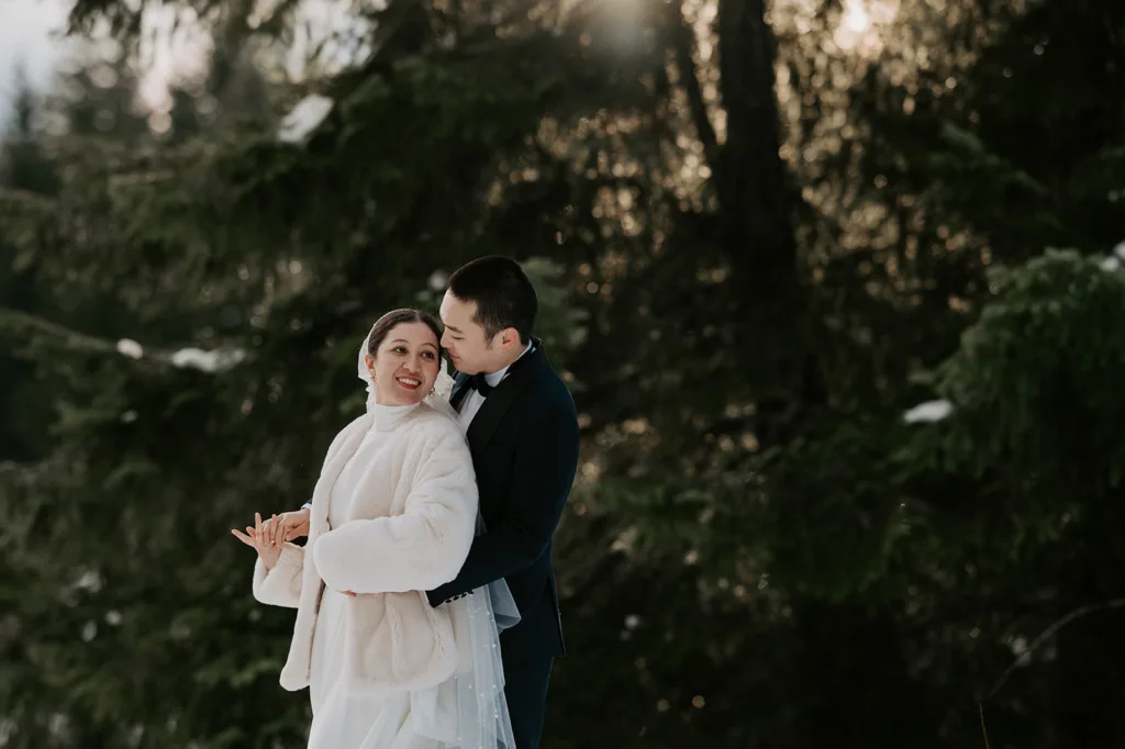 A couple in wedding attire share a tender embrace in a sunlit snowy forest, reflecting the warmth and joy of their winter elopement at Gold Creek Pond.