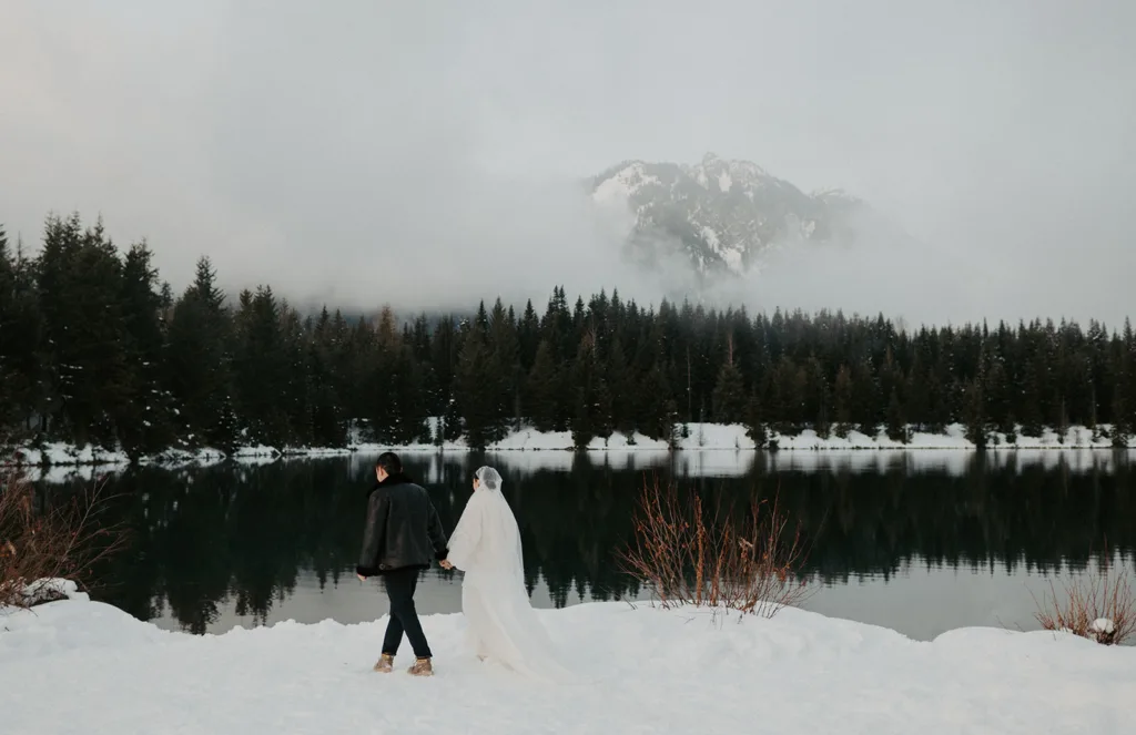 A bride in white and groom in black walk by the snowy shores of Gold Creek Pond, with pine trees and misty mountains in the background, reflecting the peace of a winter elopement.