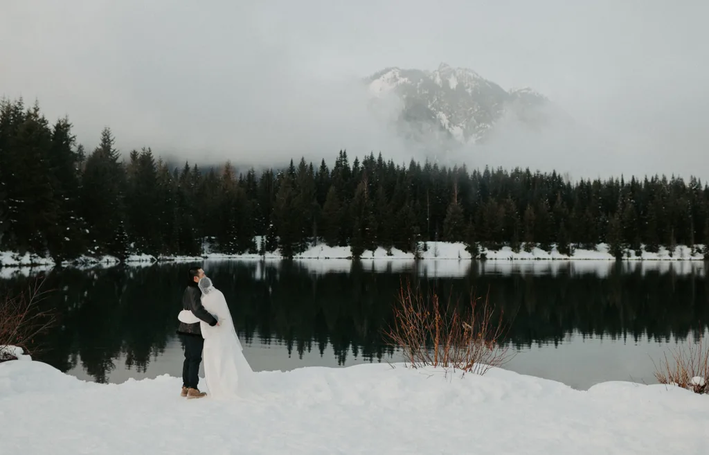 A couple stands close, enveloped in the tranquility of a snow-covered landscape at Gold Creek Pond, with forest reflections in the calm waters and a mountain shrouded in mist behind them.