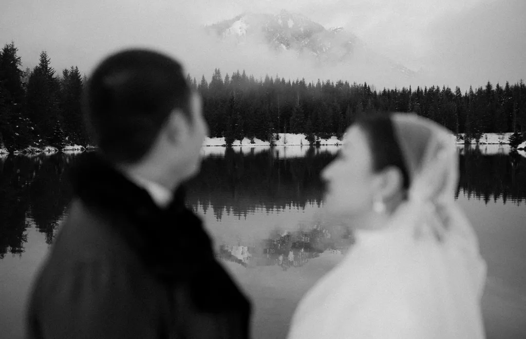 A black and white photo capturing the profiles of a bride and groom at Gold Creek Pond, reflecting the peaceful surroundings and their silent communication, encapsulated by the serene snow-dusted pines.