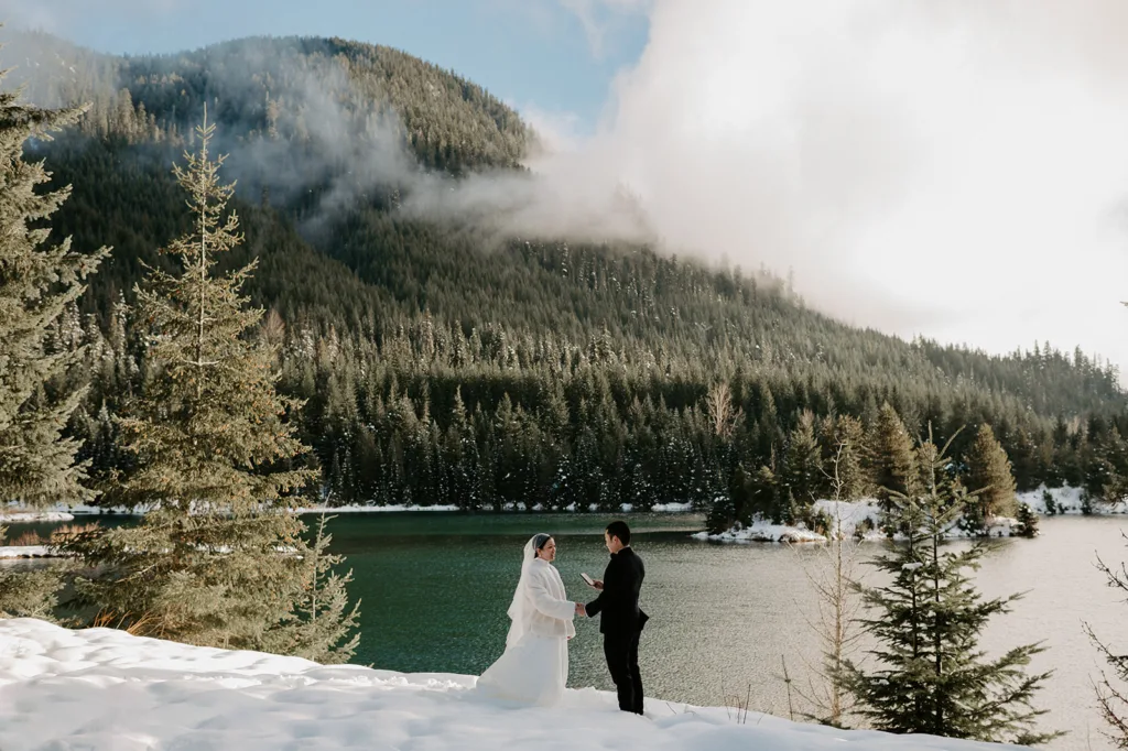 A couple stands intimately by the shimmering waters of Gold Creek Pond, with snow-dusted pines and misty mountains framing their precious elopement moment.