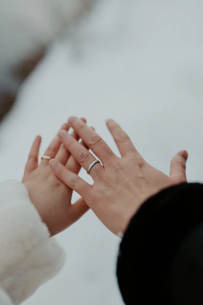 Close-up of a newlywed couple's hands with wedding rings, touching tenderly against the snowy serenity of Gold Creek Pond, symbolizing their union and commitment.