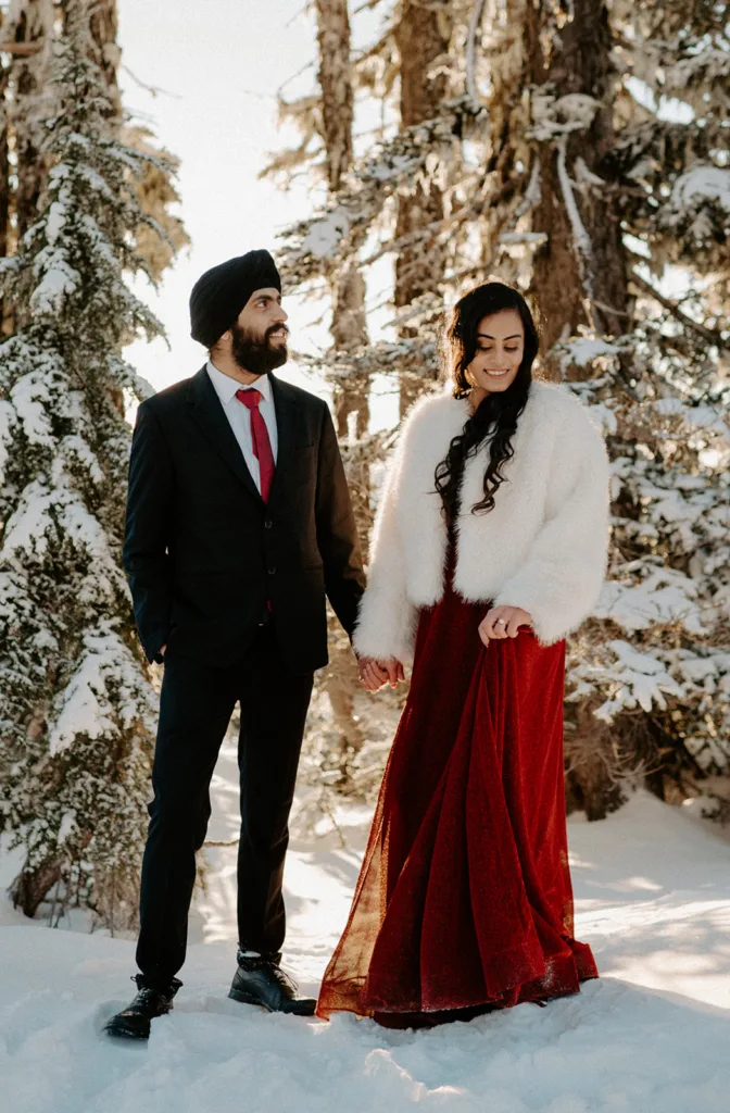 Couple walks hand in hand in a snow-laden forest, sharing a glance and a smile.