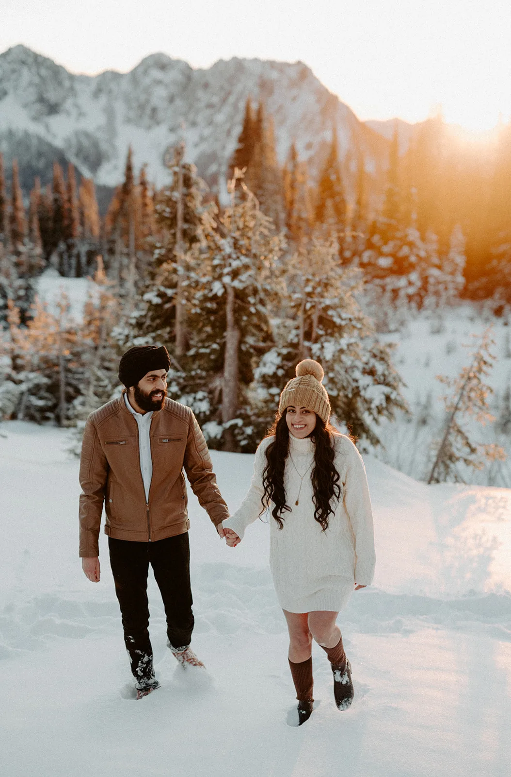 A couple holds hands, smiling as they walk through snow, with a radiant sunset over Mount Rainier National Park