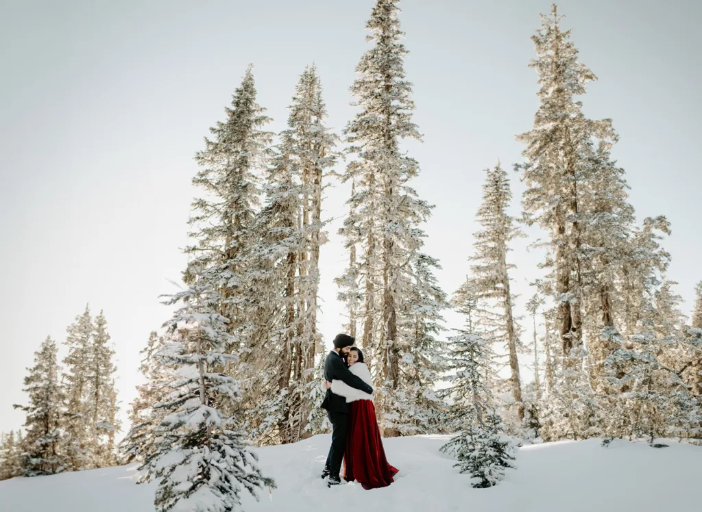 Couple embraces in a snowy forest, tall frosty trees framing the serene scene.