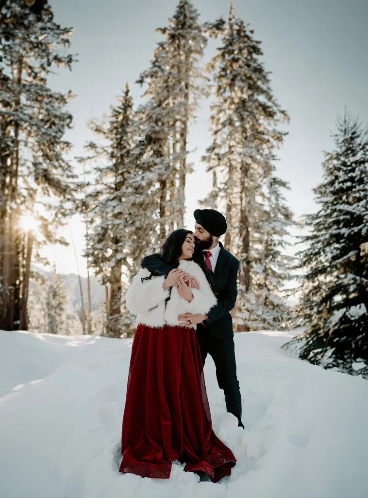 Couple kissing in a snowy forest clearing, sun peeking through the trees.