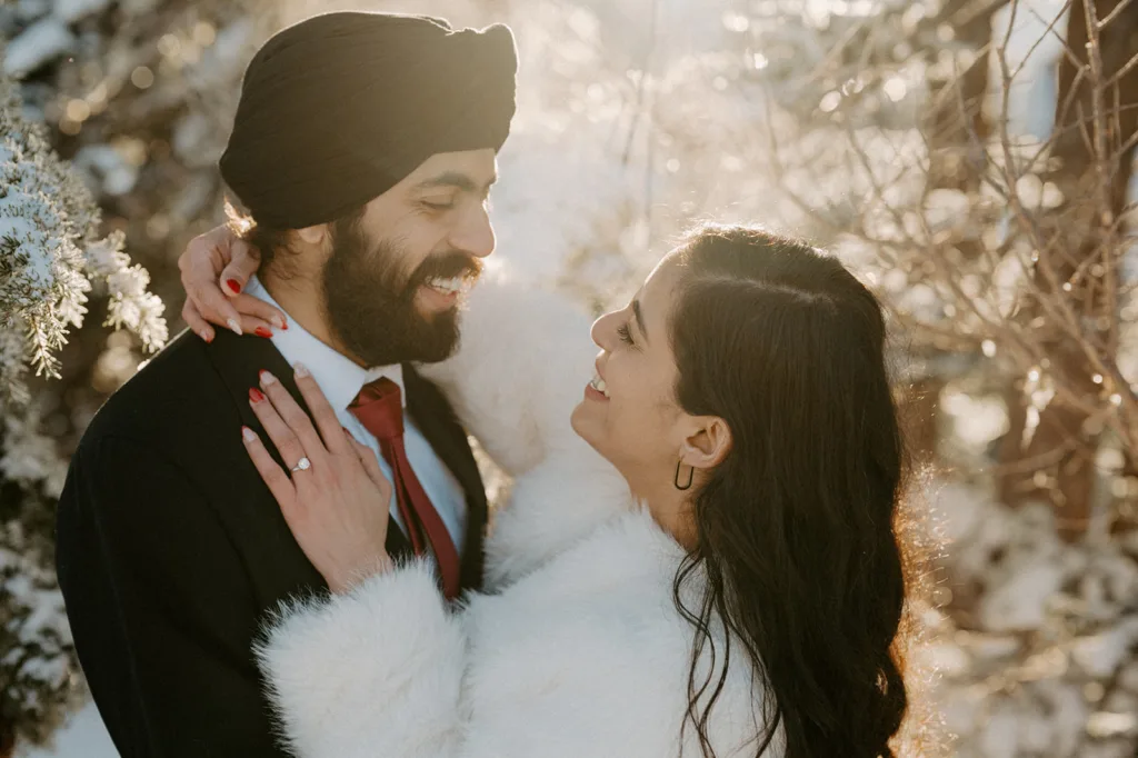 Couple smiling in a sunlit snow scene, the groom's joy reflected in his bride's gaze.