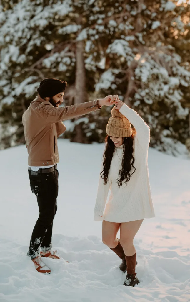 Couple shares a playful twirl in snowy woods, with warm sunlight filtering through the trees.