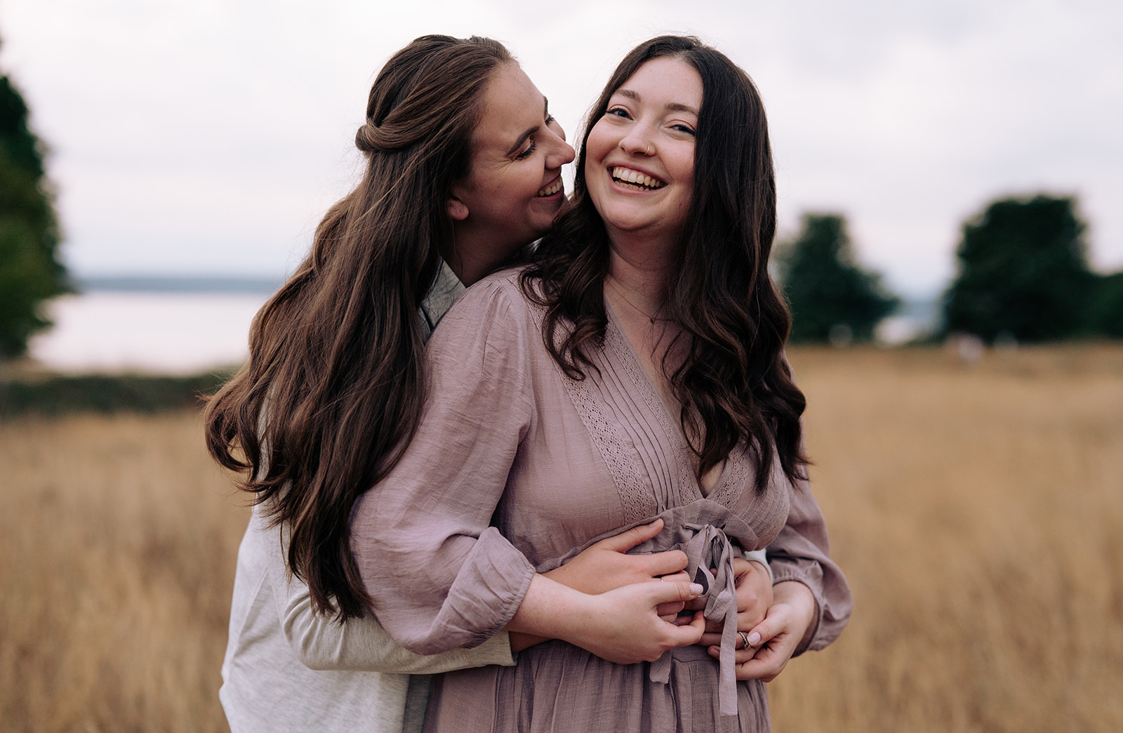 A happy couple embracing in a field, both smiling and sharing a joyful moment, with the woman in a lavender dress and the other in a light gray sweater.