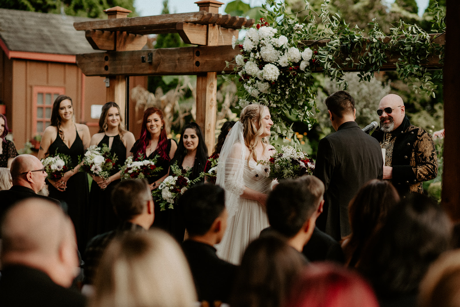 Bride standing at the altar with bridesmaids in black dresses beneath a wooden pergola adorned with white florals at Willows Lodge