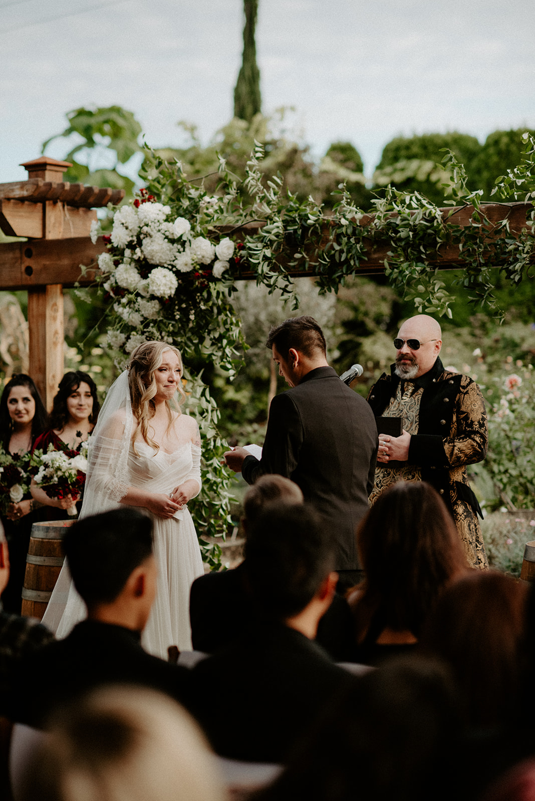 Wide ceremony shot of bride and groom exchanging vows under a floral pergola arch at Willows Lodge with guests seated around them