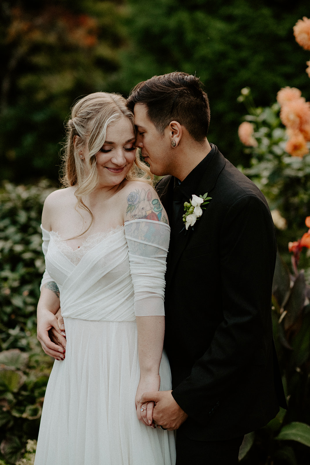 Groom kissing his bride's cheek as she smiles, dahlias blooming in the garden behind them