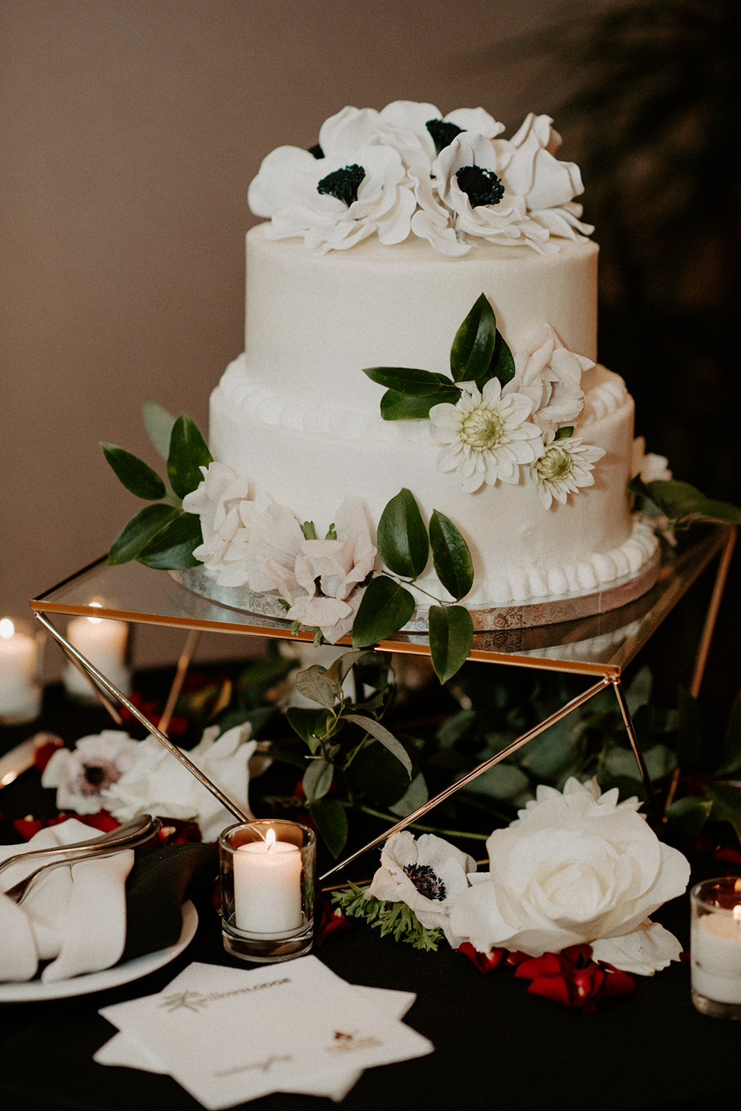 A two-tier white wedding cake decorated with anemone flowers and greenery, flanked by candles