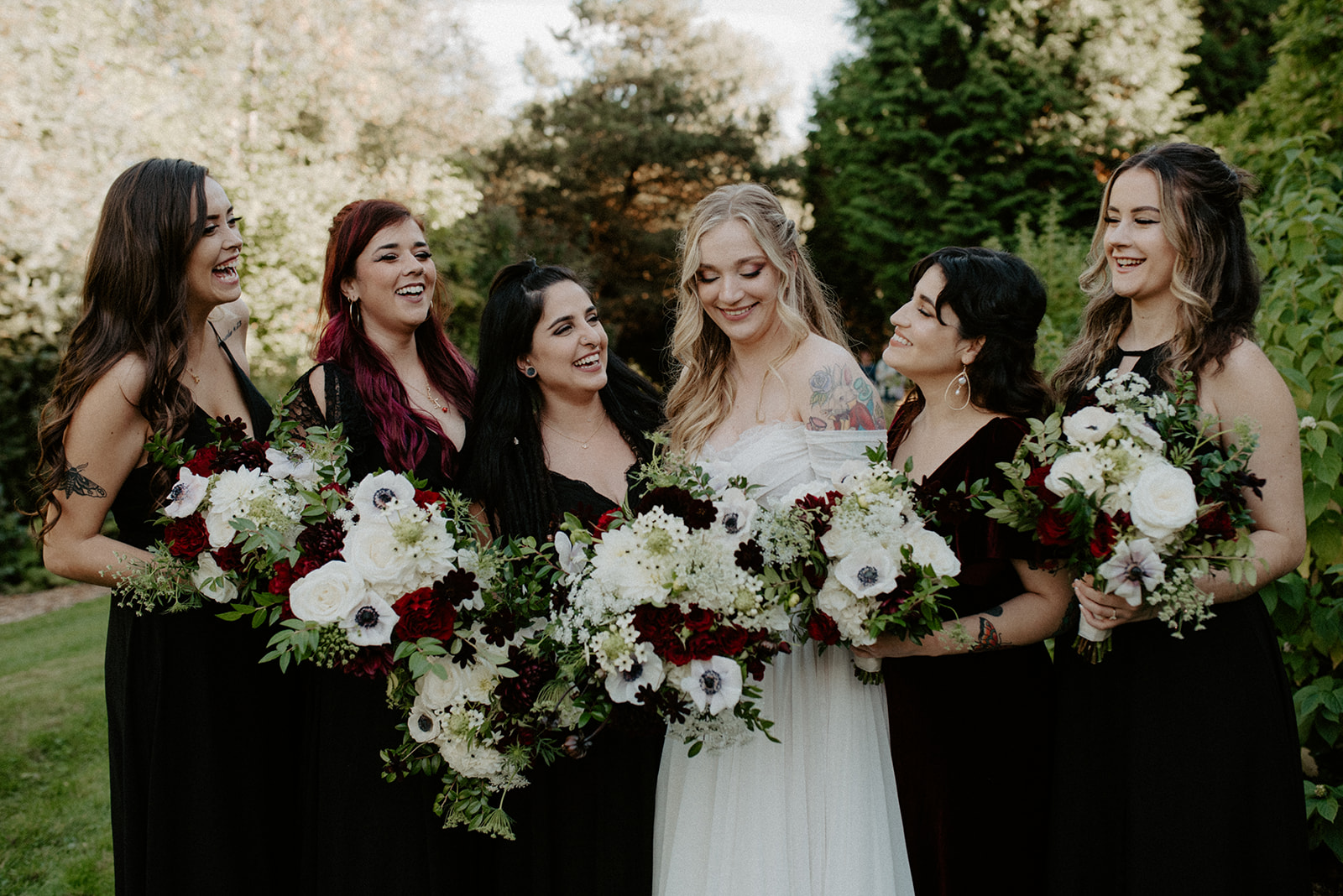 Bride with her bridesmaids in black dresses, all laughing together while holding dark red and white bouquets in the Willows Lodge garden