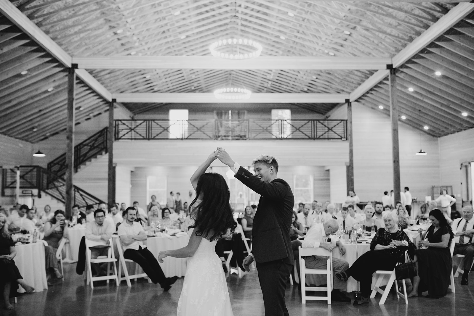 A black and white photo of a bride and groom sharing their first dance in a rustic venue, with the groom spinning the bride while guests look on and smile, capturing the joyous atmosphere.