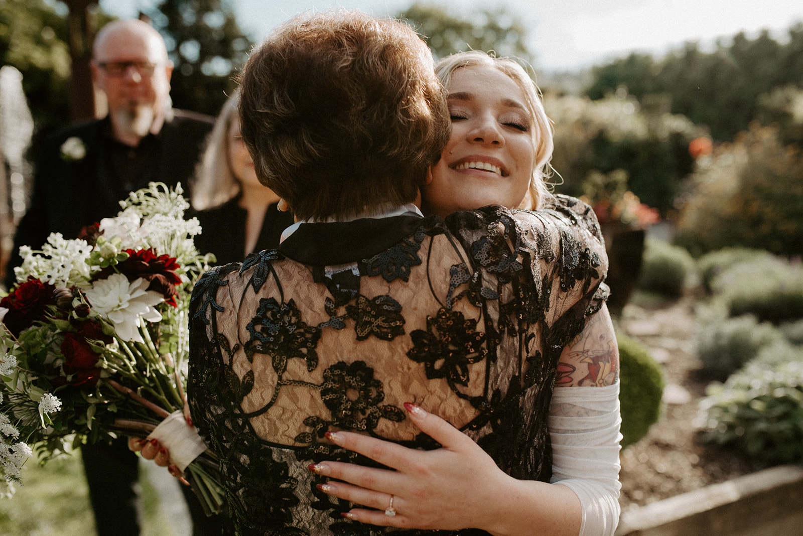 Bride embracing a family member in the garden before the ceremony, holding a bouquet of dark red and white flowers