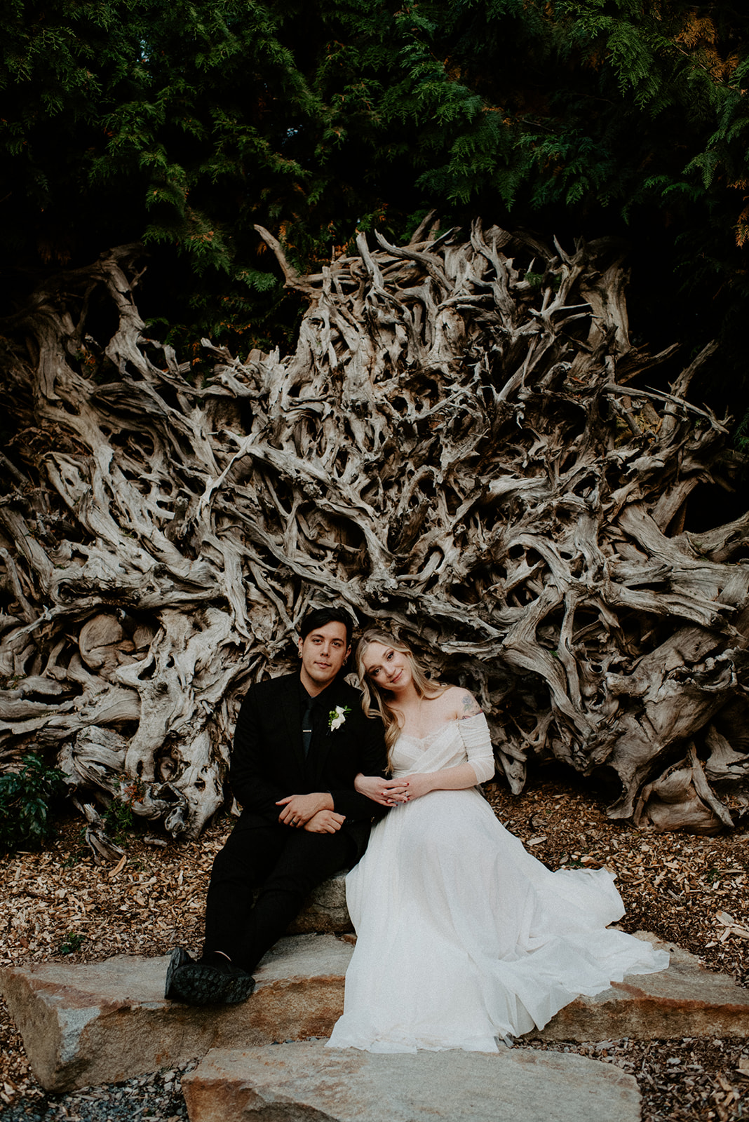 A couple sitting in front of an intricate driftwood wall, with the woman in a white dress leaning on the man in a dark suit, both looking serene and content.