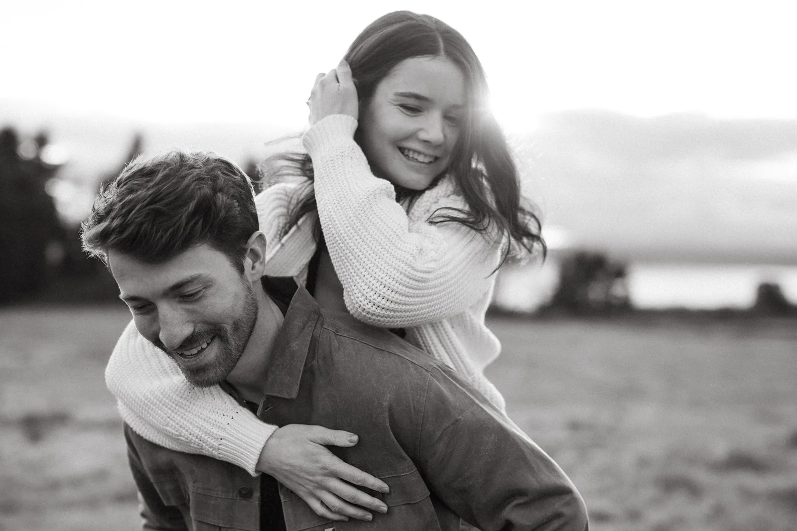 A black and white photo of a couple sharing a playful piggyback ride in a field, with the woman laughing and holding on to the man's back.