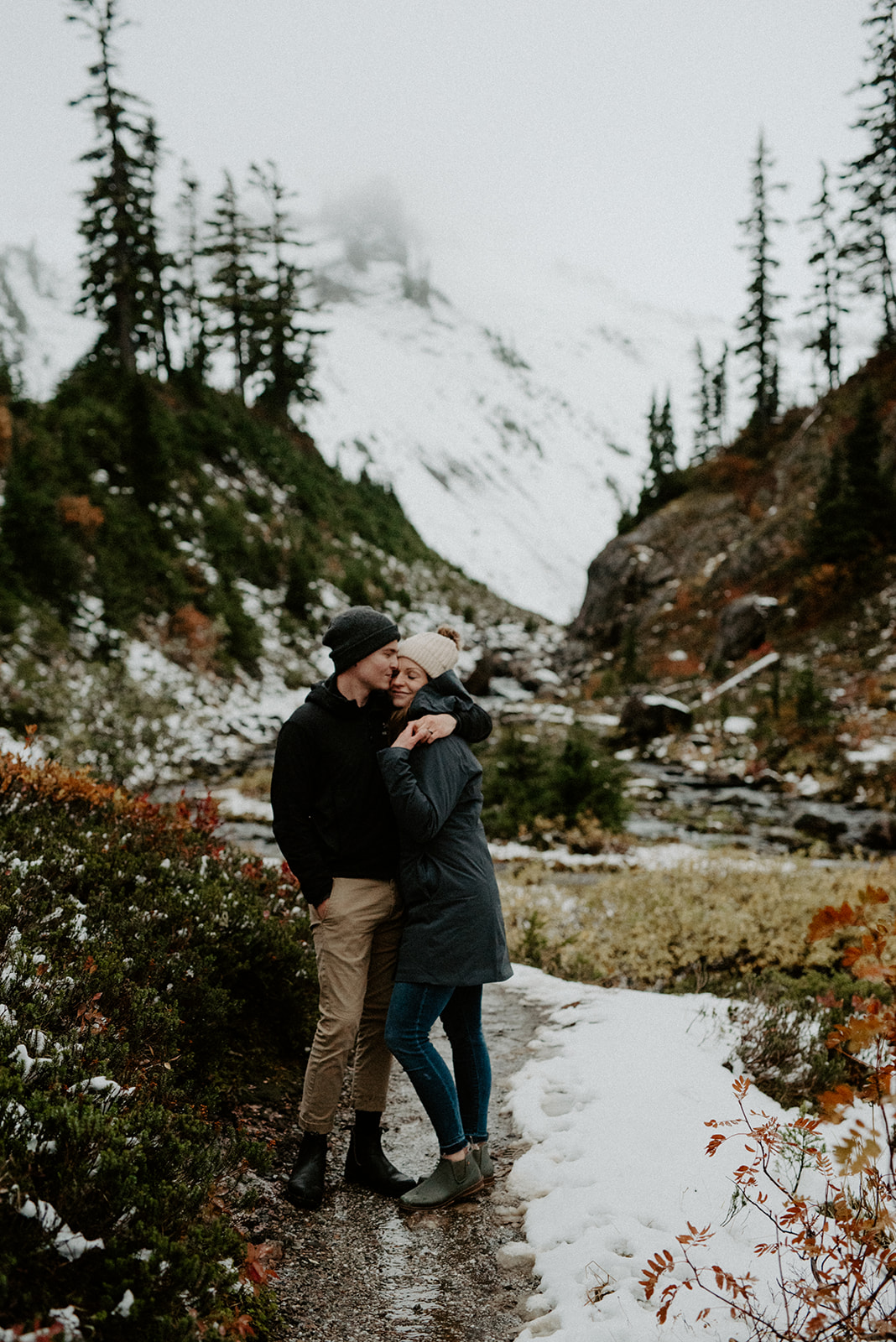 A couple shares a loving embrace during a winter stroll in the mountains, surrounded by snow-covered paths, evergreen trees, and rocky terrain.