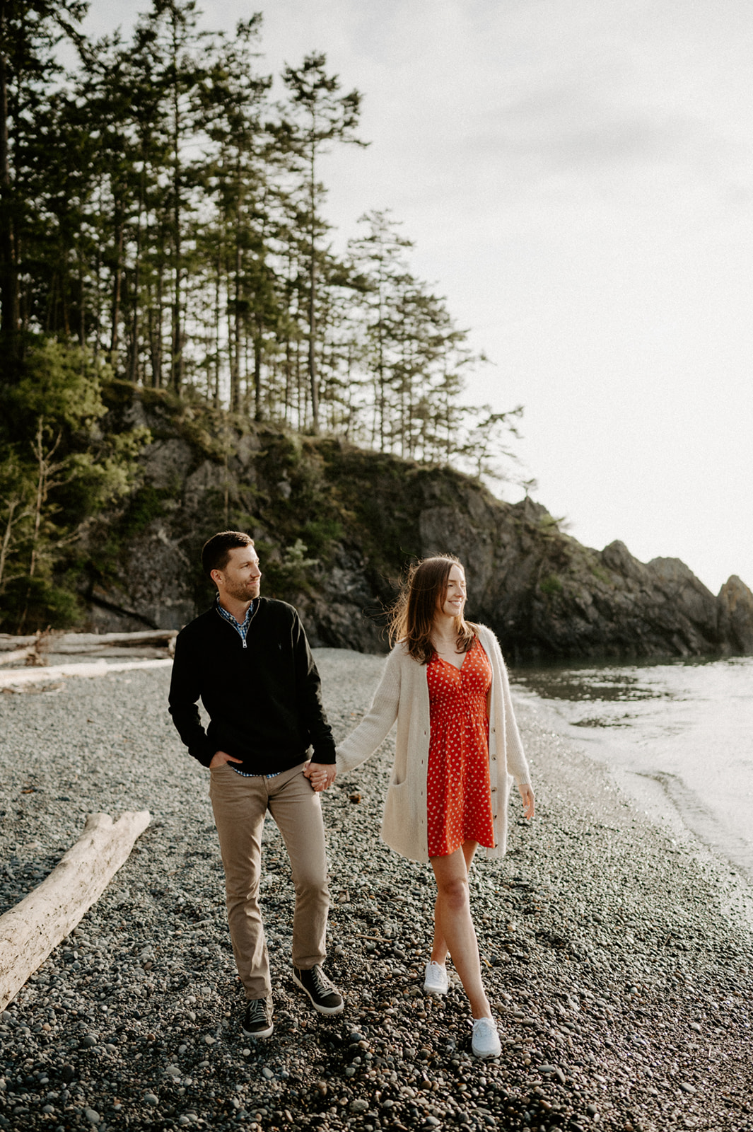 A couple walking hand in hand along a rocky beach, surrounded by trees and calm water.