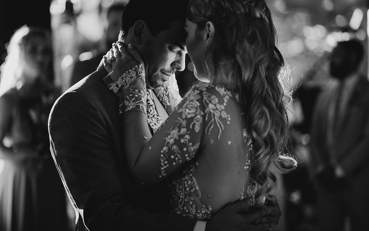 A black and white photo of a bride and groom sharing their first dance, embracing closely with the bride's detailed lace dress and the groom's tender expression highlighting the emotional moment.