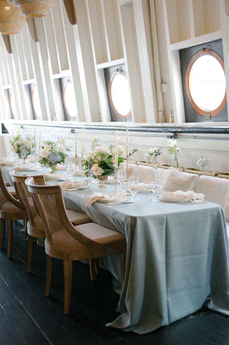 A refined dining area on The MV Skansonia, showcasing a long table with a blue tablecloth, white floral arrangements, and tall candlesticks, surrounded by wooden chairs and porthole windows.