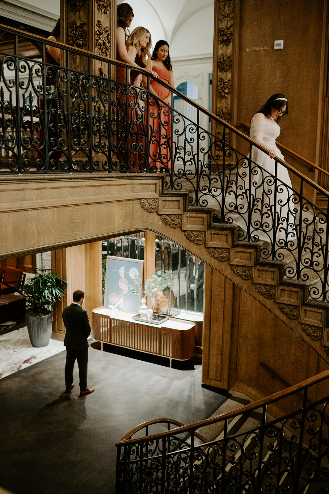 Bride descends an ornate staircase while bridesmaids look on and groom waits below at the Fairmont Olympic Hotel.