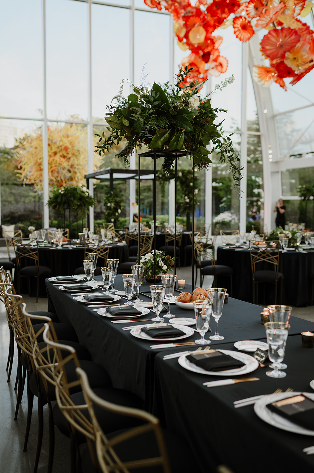 Table settings at Chihuly Garden and Glass with black tablecloths, gold-rimmed glasses, greenery centerpieces, under a vibrant glass flower ceiling installation.