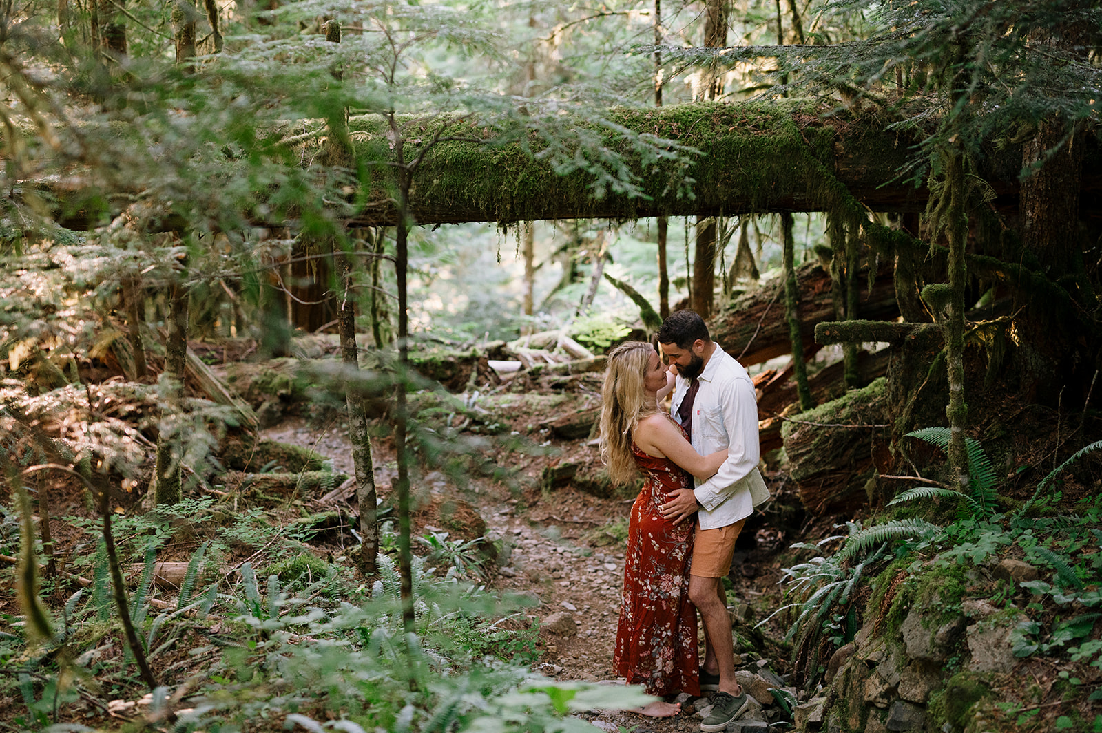 R and M standing together under a moss-covered log in a forest.