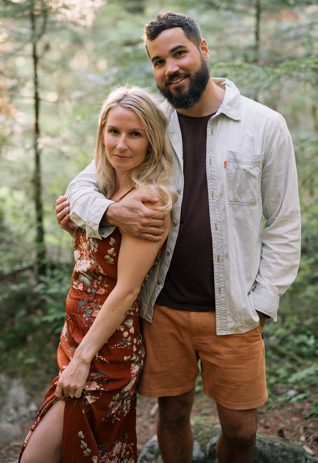 R and M smiling together in a forest during their engagement session.