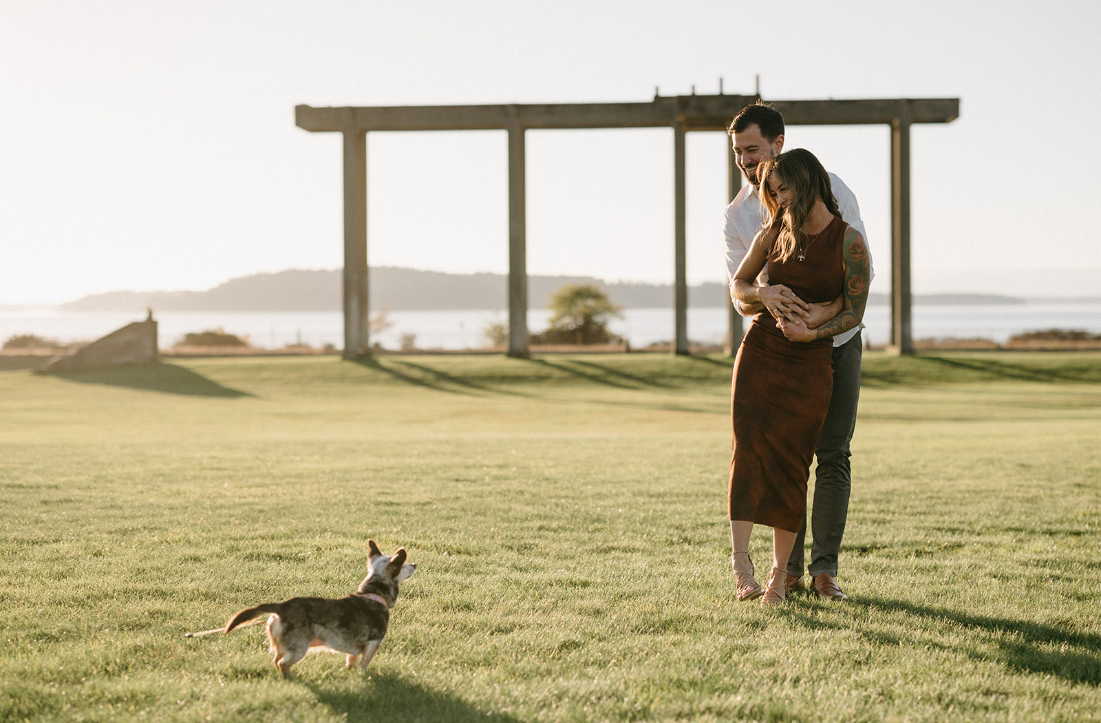 A couple embracing while their chihuahua plays on the grass.