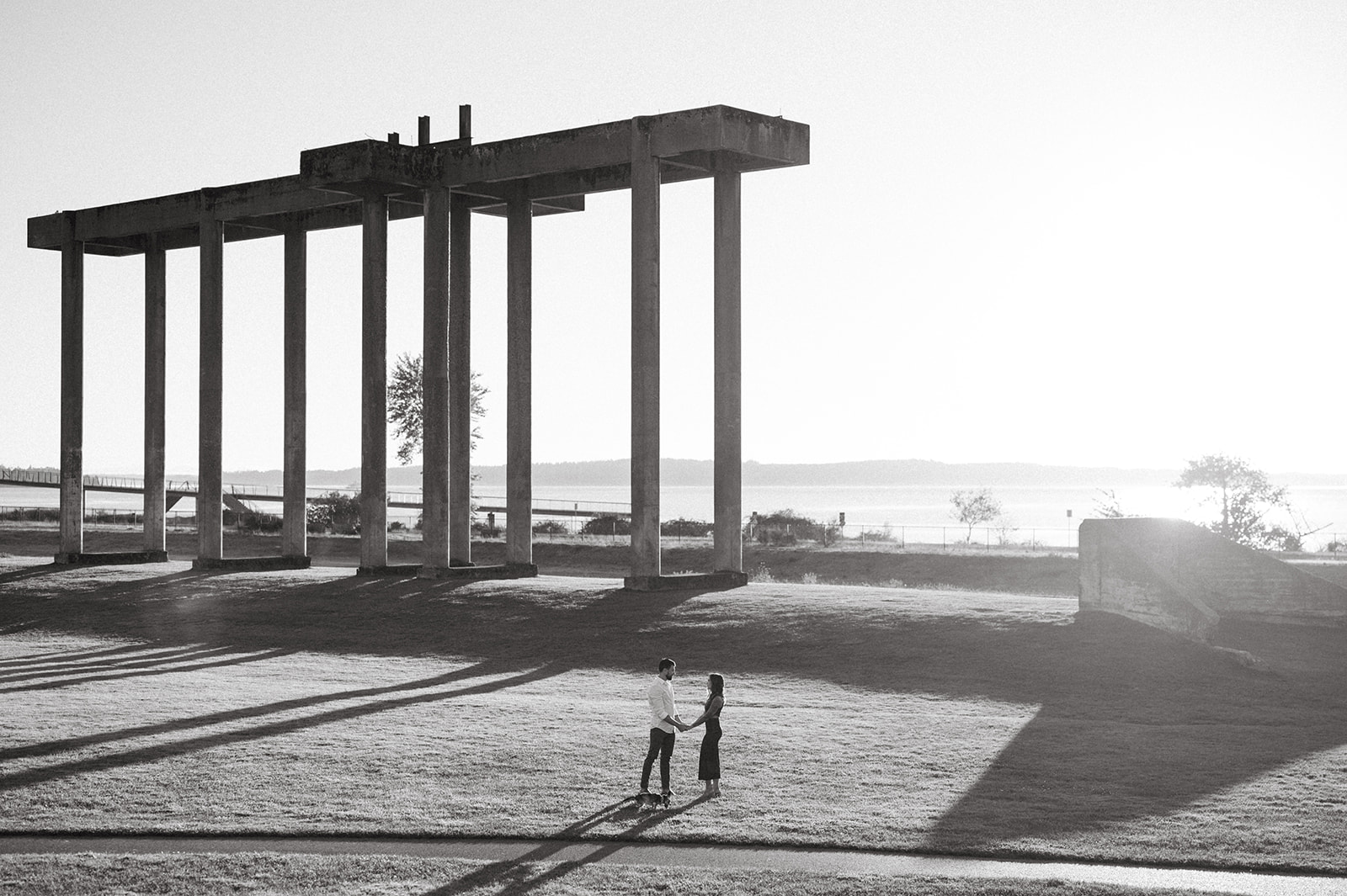 A couple holding hands under a large concrete structure by the water.
