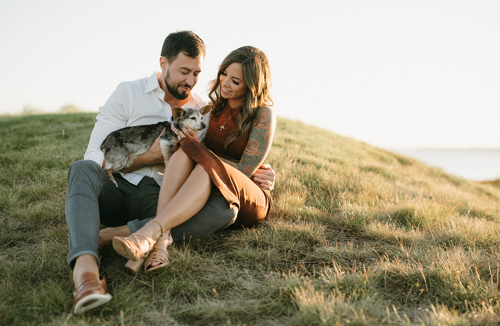 A couple sitting on a grassy hill with their chihuahua.