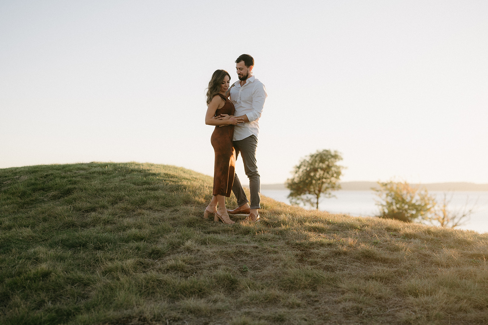 A couple standing closely on a grassy hill at sunset.