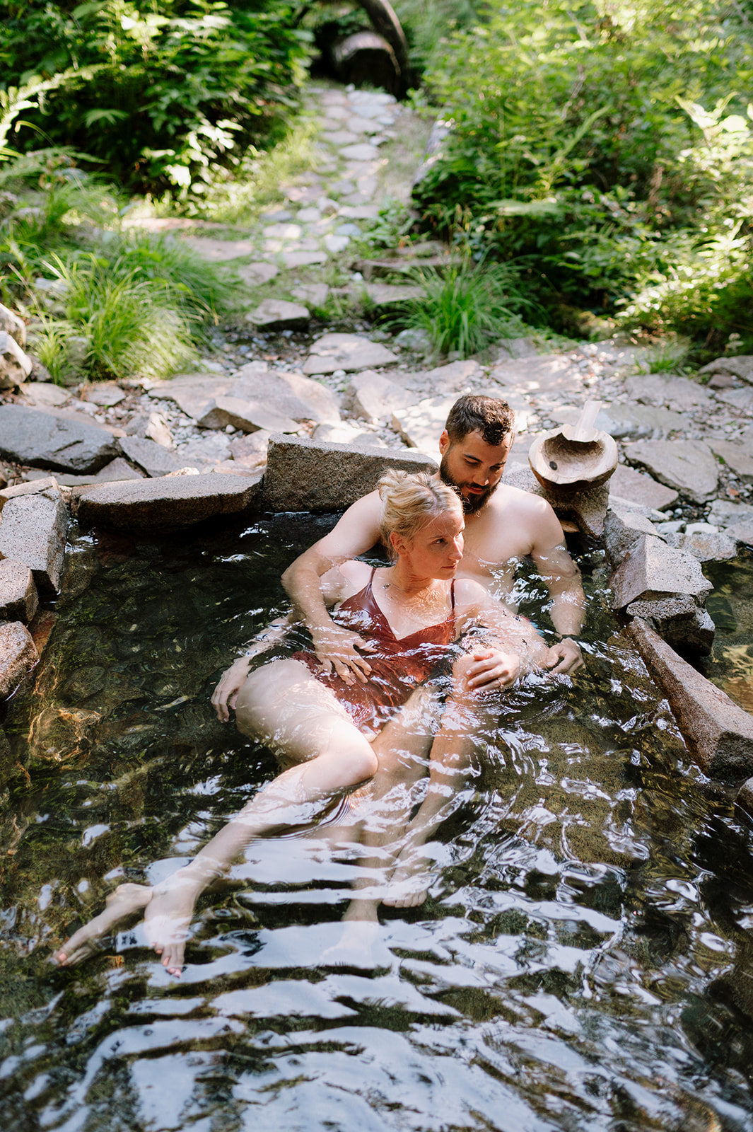 R and M lounging together in a natural hot spring surrounded by greenery.